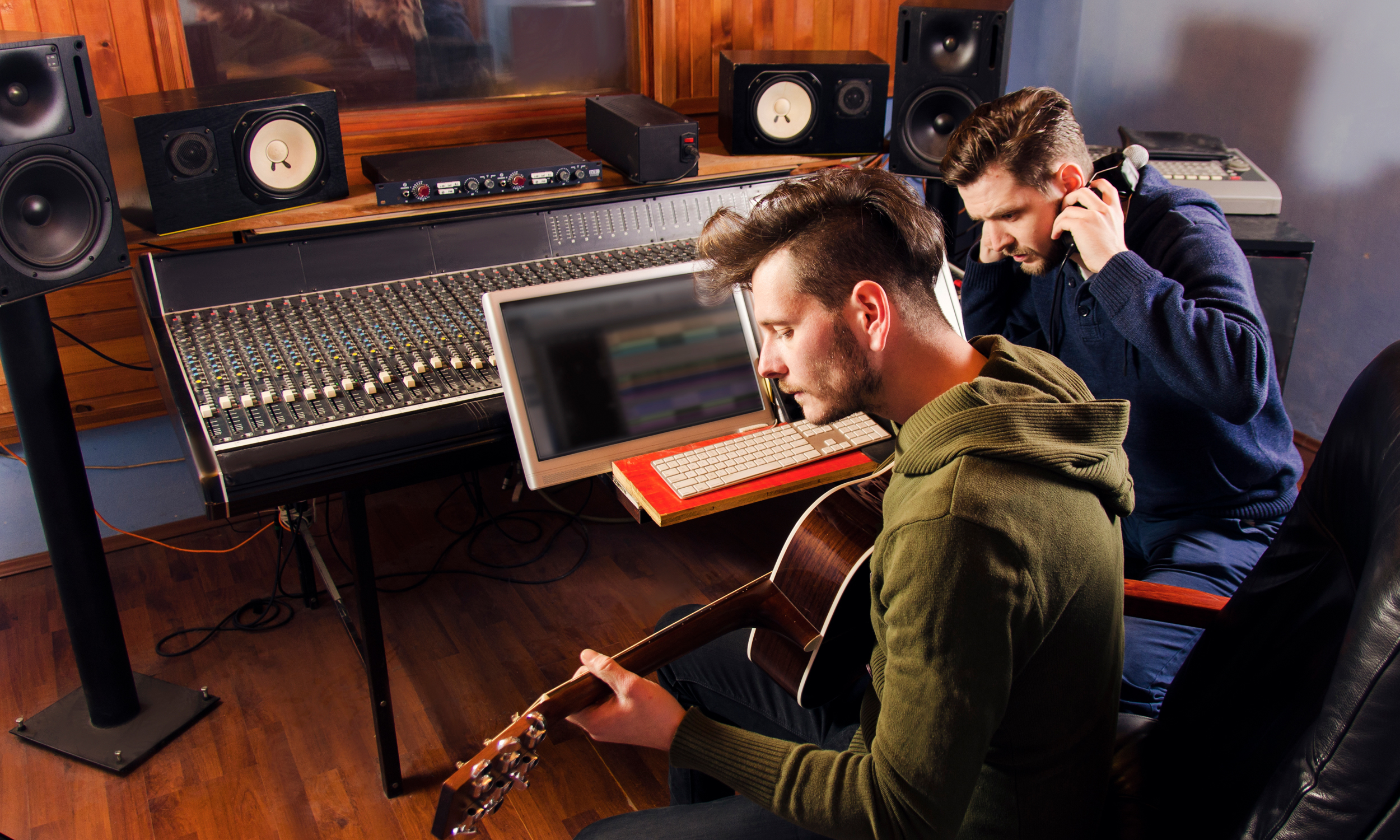 Audio studio with two men performing. One man has a guitar and the other is listening in a pair of headsets.